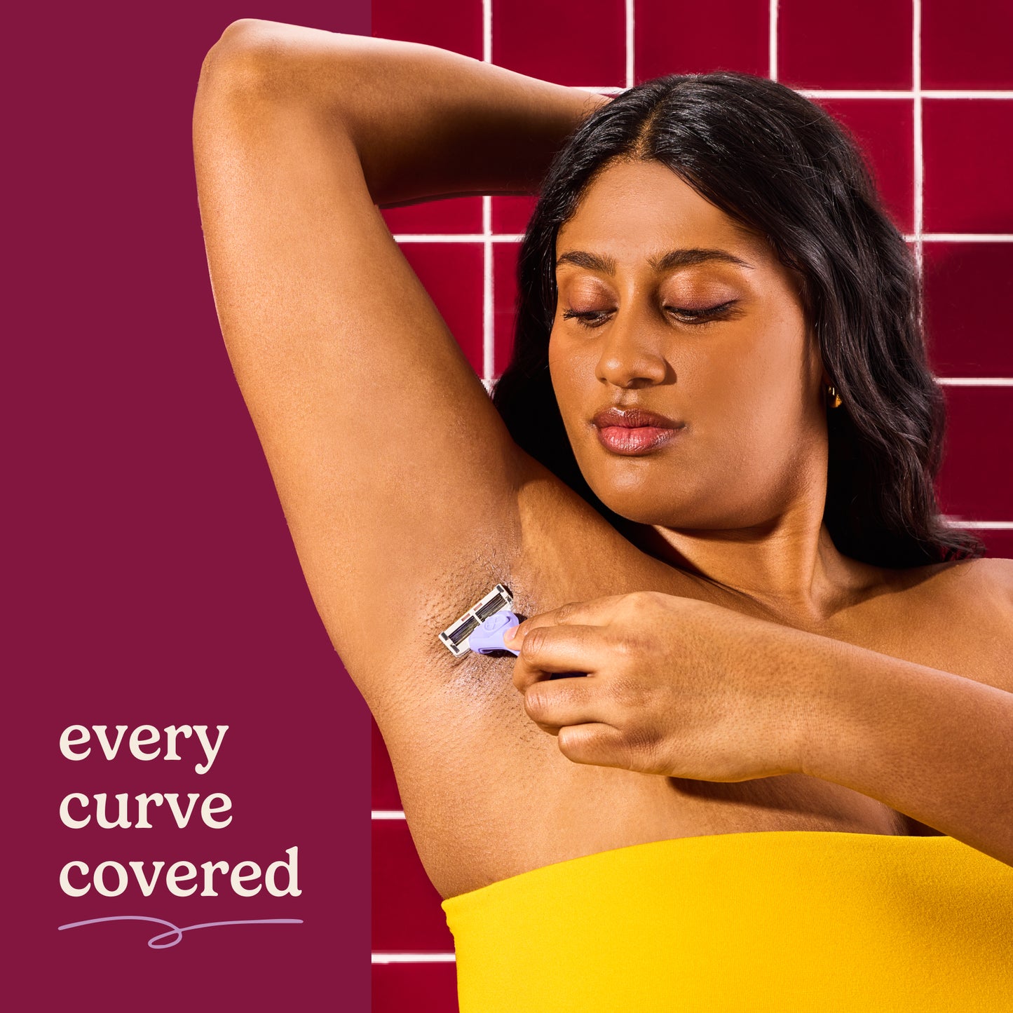 Woman shaving her arm with a razor, wrapped in a yellow towel, against a red tiled wall.