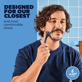 Man shaving with a razor, wearing a blue shirt, against a blue tiled wall background.