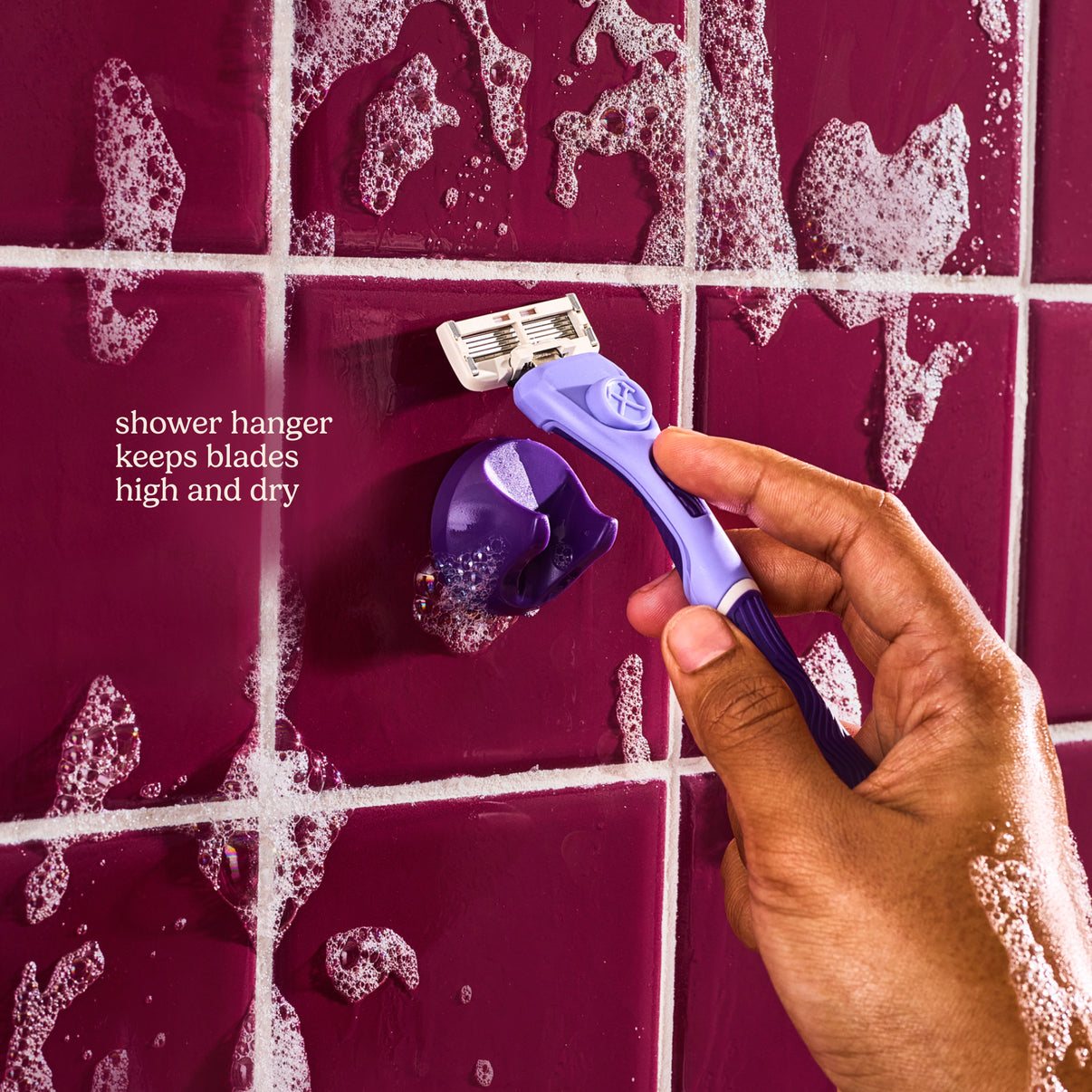 Hand holding a purple razor against a tiled shower wall with razor hanger.