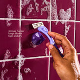Hand holding a purple razor against a tiled shower wall with razor hanger.