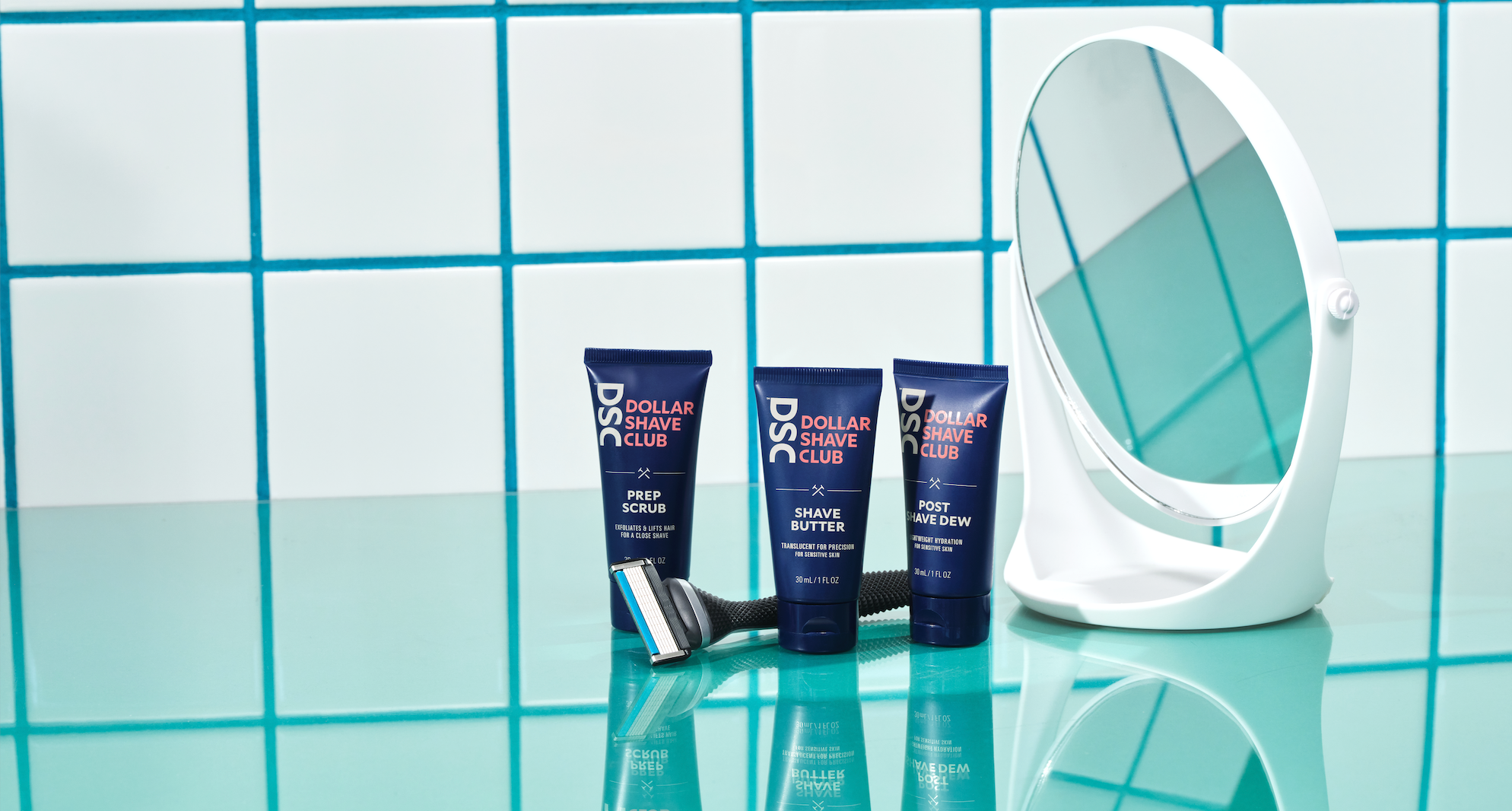 Dollar Shave Club products and vanity mirror sitting on aqua colored surface in front of a tiled wall.