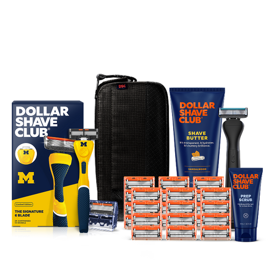 Dollar Shave Club University of Michigan grooming kit on a white background. Includes a navy razor handle with the Michigan Wolverines "M" logo, 24 blades, Shave Butter, Prep Scrub, a black 3-in-1 electric trimmer, and a black traveler case.